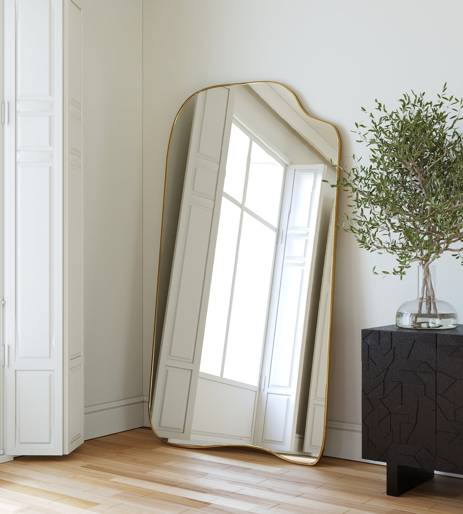 Asymmetrical full-length mirror with an organic shape and thin brass-colored frame, leaning against a wall in a bright room with tall windows, light wood flooring, and a potted plant.